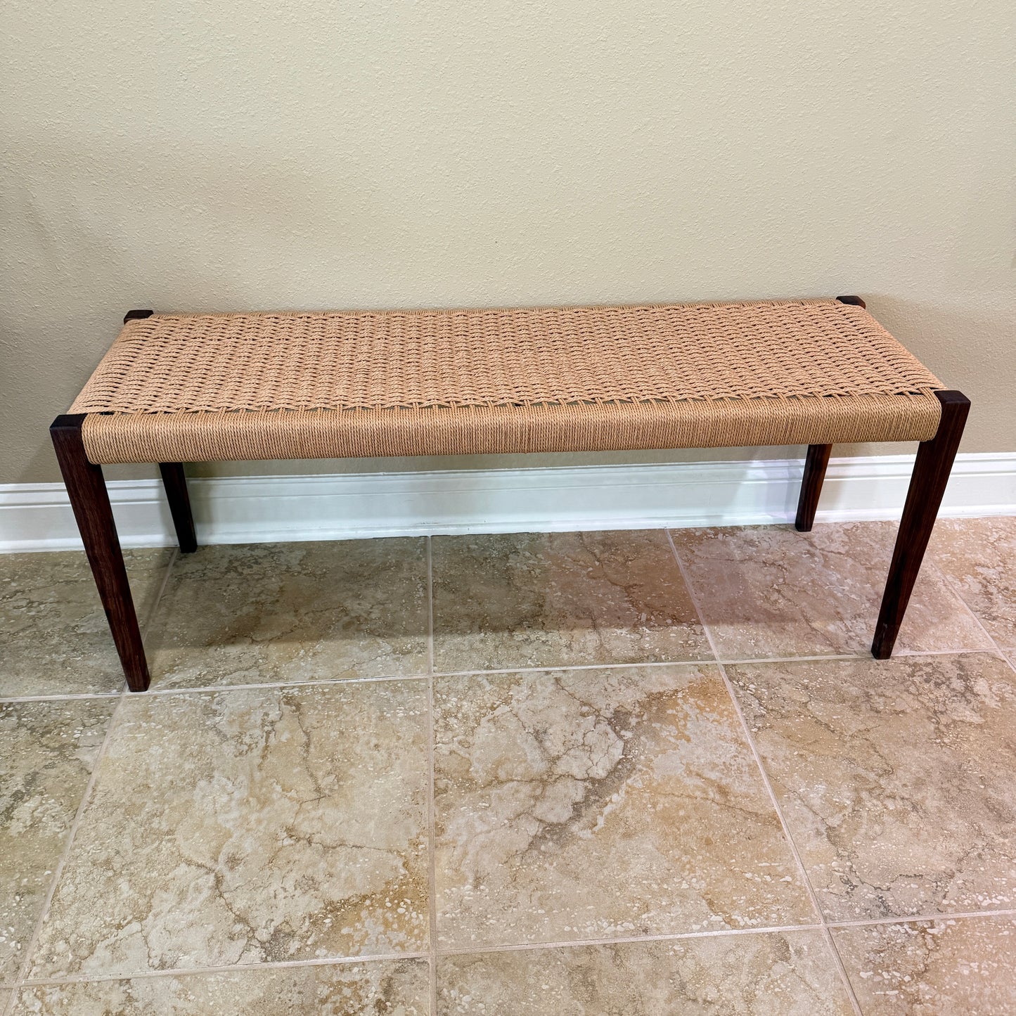 Side view of bench with tapered walnut legs and a woven seating area sitting against a wall on a tile floor.