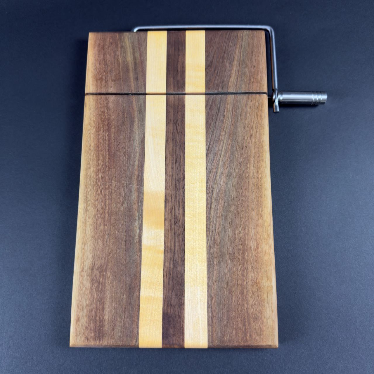 Cheese cutting board shown from and end view from the top. Chrome finish cutting handle and arm. Colorful linear pattern of walnut and maple. Measure a generous 8 x 13. Extra cutting wires included. Please contact me for additional assistance.