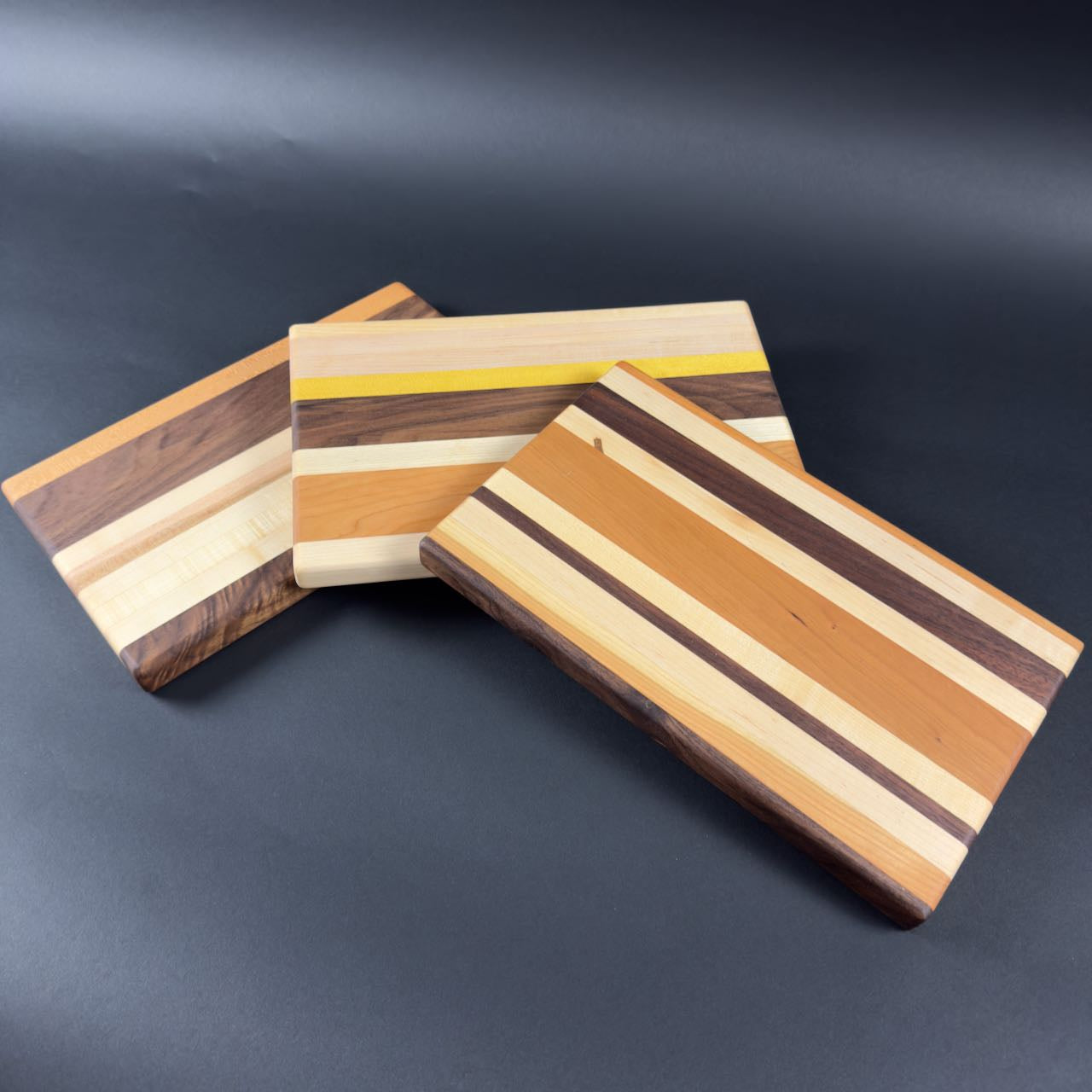 Three compact hardwood chopping boards with striped patterns on a dark surface. Showing each with different patterns.