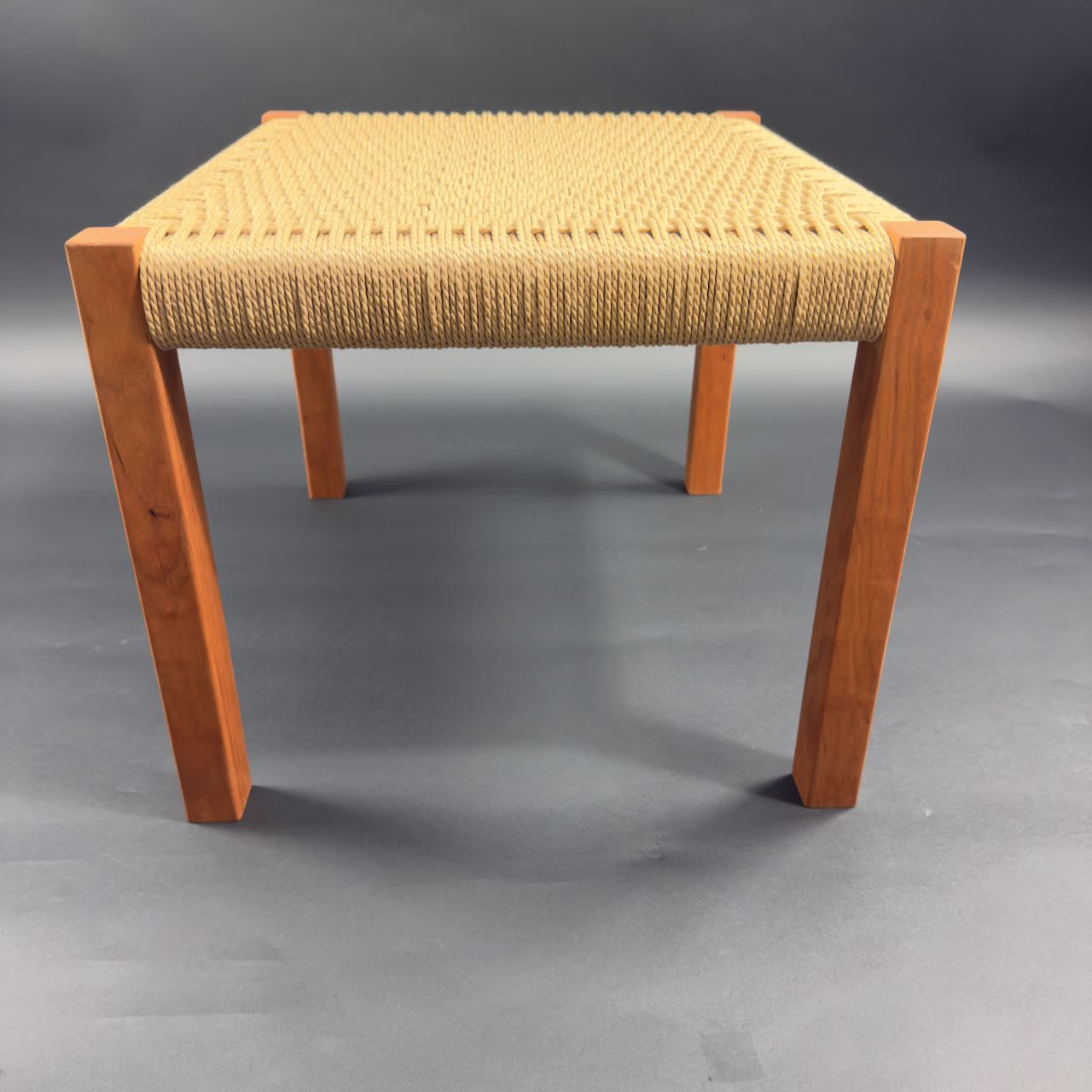 Beautiful  square cherry wood foot stool with danish cord weave seating, side view on a dark background. 16 x 16 and 17 inches high