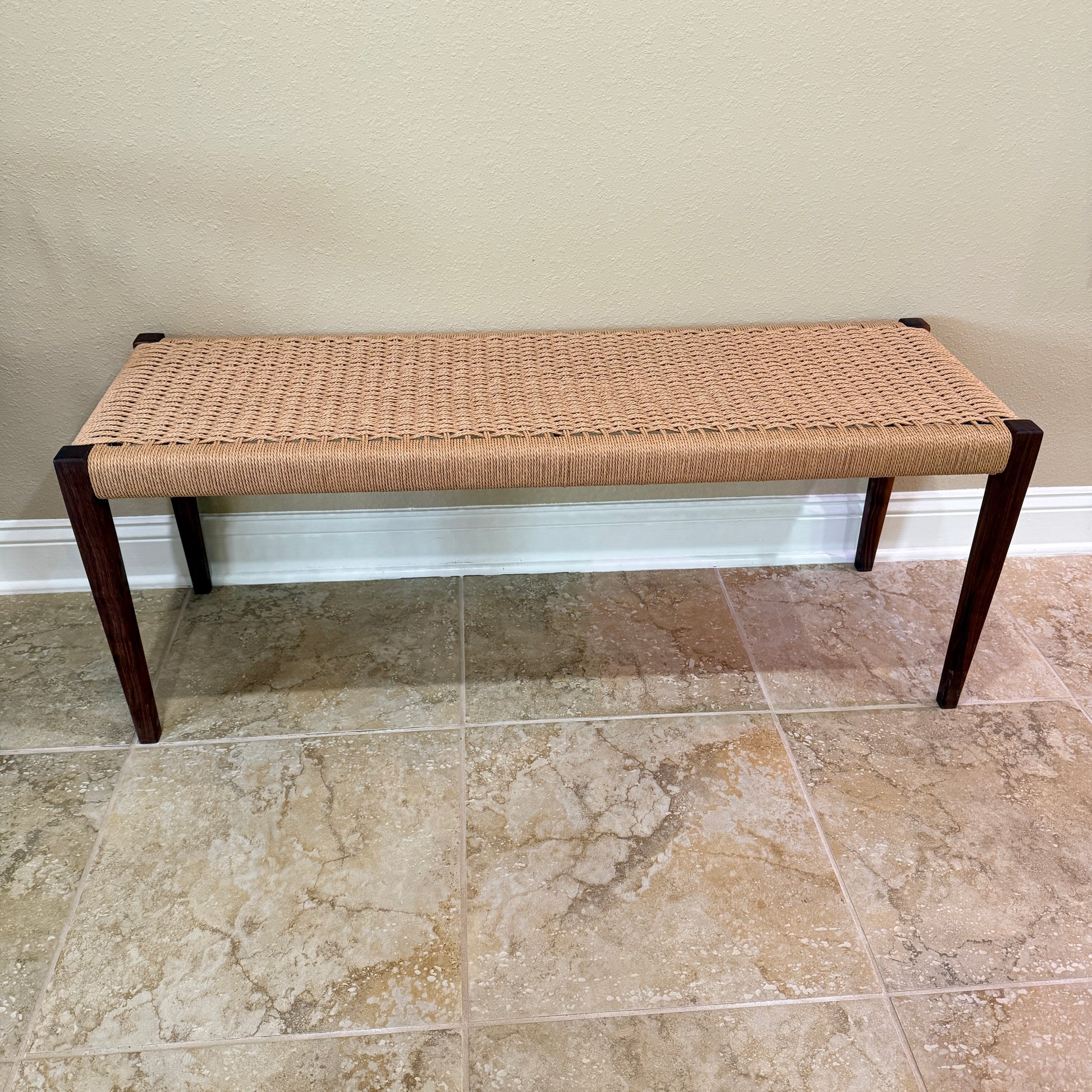 Side view of bench with tapered walnut legs and a woven seating area sitting against a wall on a tile floor.