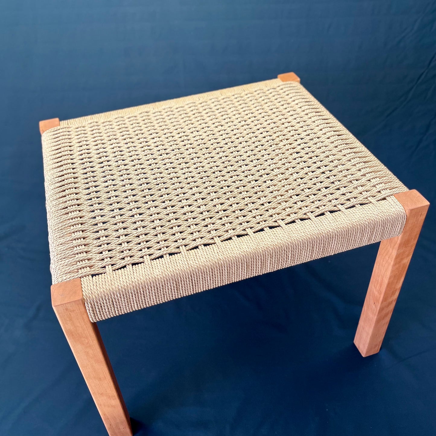Beautiful  square cherry wood foot stool with danish cord weave seating, angle view on a dark background. 16 x 16 and 17 inches high