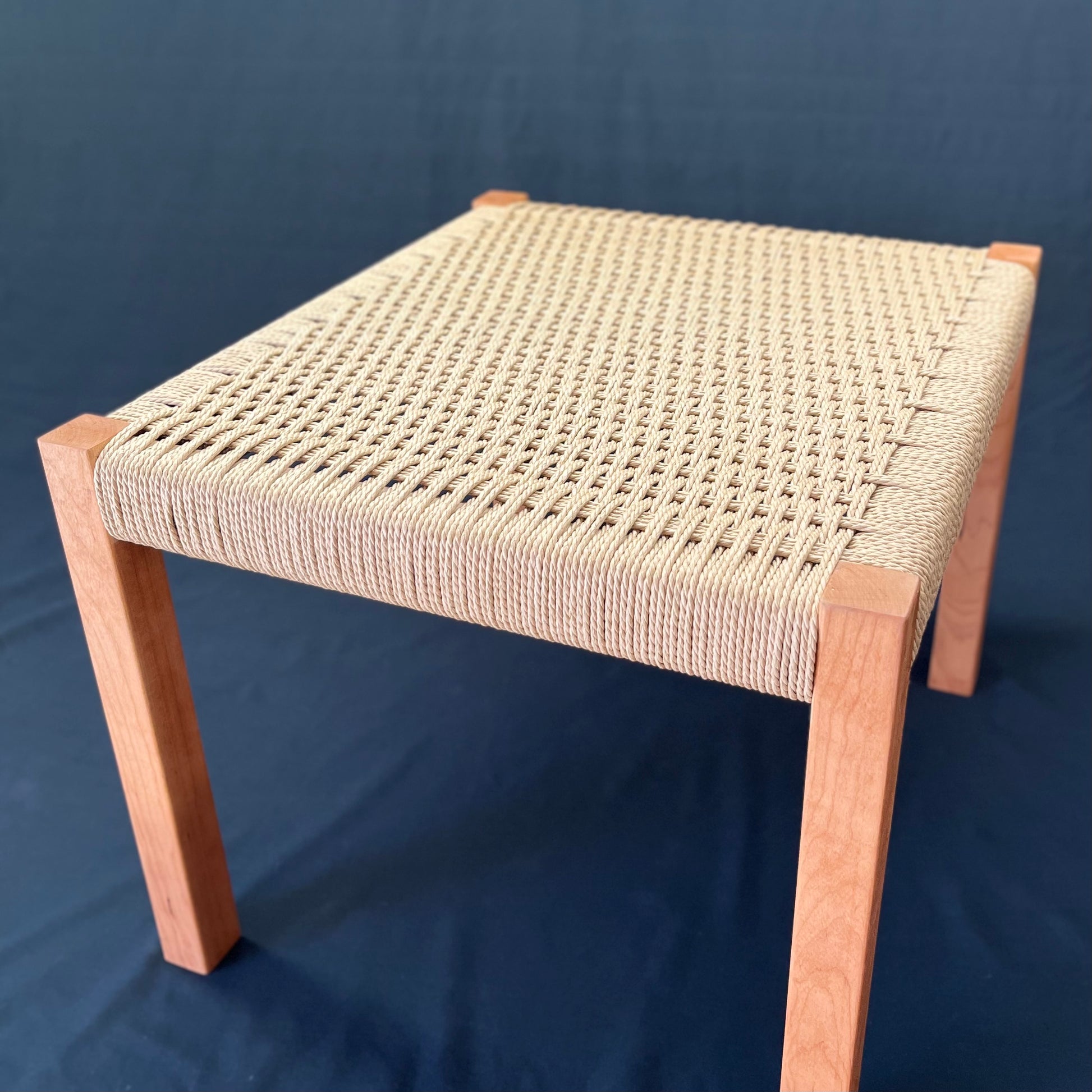 Beautiful cherry wood foot stool with danish cord weave seating angle view on a dark background.